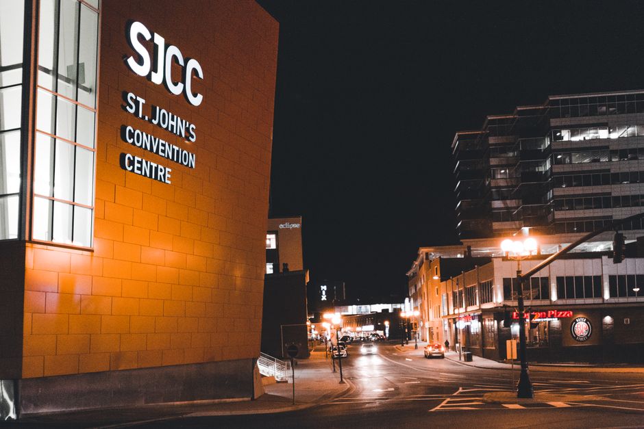 A nighttime scene outside St. John's Convention Centre (SJCC), with the building's orange facade illuminated by exterior lighting, displaying the sign 'SJCC St. John's Convention Centre' on the wall. To the right, a street view reveals a wide road with parked cars and a few moving vehicles under streetlights. Adjacent to the convention centre, there are commercial buildings, including a pizza restaurant and other shops, with illuminated signage and glass windows. The scene also shows a traffic light and street lamps casting a warm glow on the pavement and a few pedestrians. This setting captures the urban environment where local house removals or furniture transport may be scheduled, with the surrounding infrastructure and lighting typical of a bustling city area, suitable for coordinating transport and packing logistics related to home relocation projects as coordinated by Man with Van St Johns.
