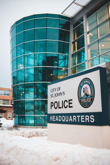 Exterior view of the City of St. John's Police Headquarters, featuring a modern curved glass building with predominantly blue-tinted windows and metallic framing. In the foreground, a large white sign displays the police department's emblem and lettering, indicating 'City of St. John's Police Headquarters.' The ground is covered with snow, suggesting winter conditions. The image is taken during daytime with overcast skies, and part of a neighboring multi-story building is visible on the left side. This setting illustrates the location involved in house removals and relocation logistics, with the police headquarters serving as a landmark in St. John's, supporting the need for road closures related to home moving activities organized by local services such as Man with Van St Johns.