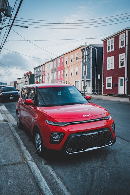 A red Kia Soul compact car parked partially on the pavement and street of a residential area during daylight hours, with multicoloured modern terraced houses lining the background. The car is positioned close to a curb, with its front facing the camera, showcasing the distinctive grille and headlights. Overhead, multiple power lines cross the sky, which is partly cloudy with patches of blue. The street appears quiet and is situated in a neighbourhood suitable for home relocation and furniture transport. The image illustrates typical urban parking conditions where managed parking and careful loading are essential for house removals, as supported by services like Man with Van St Johns. The scene depicts the environment involved in planning efficient moving logistics, including parking considerations, loading processes, and residential access for professional removals.