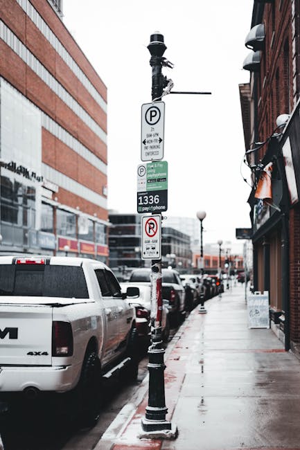 A street scene showing multiple parked vehicles along a pavement, including a white van positioned close to the camera with its rear end visible. Adjacent to the vehicles, a metal parking bay pole is equipped with multiple parking and regulatory signs, including a parking zone sign, pay-by-phone parking information, and a no-parking restriction. The pole is secured with a black base and has a mounted streetlight on top. On the right side of the image, there are brick buildings with storefronts, some featuring awnings and street signage. The pavement is wet, indicating recent rain, and the environment suggests an urban area suitable for home relocation or furniture transport activities, as supported by the signage that assists in avoiding parking fines during removals or moving services. The image reflects an organised street setup for logistical planning related to house removals, with proper parking management for moving companies like Man with Van St Johns.
