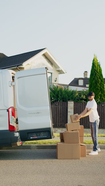 A white panel van belonging to Man with Van St Johns is parked on a cobblestone driveway in front of a residential property with a red roof and white picket fence. Inside the vehicle, a mover wearing 