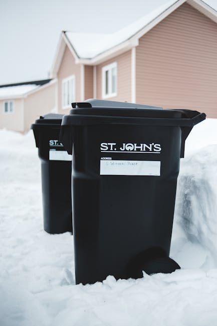 Two black wheelie bins with white labels, marked for waste disposal, are positioned outside a residential property during winter with snow covering the ground. One bin, in the foreground, has a label indicating 'S. St. Johns, Waste Paper,' while the second bin, slightly behind and to the left, has a partially visible label. The bins are situated near a paved pathway leading to the house, which has a light pink exterior with white window frames and a sloped roof. The scene captures the process of waste collection following a home declutter or house relocation, with the bins awaiting emptying or disposal. The environment is well-lit with natural daylight, and the residential setting reflects typical elements involved in house removals and packing logistics. Man with Van St Johns provides services related to this type of home clearance, ensuring efficient waste management during relocations or property clear-outs.