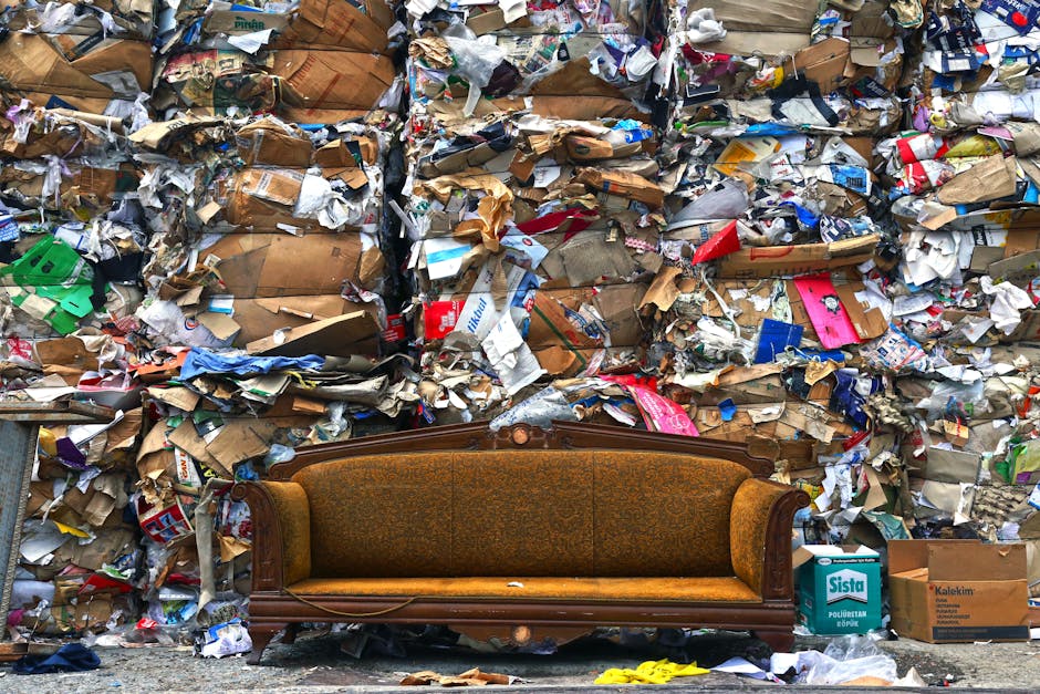 A vintage upholstered sofa with a wooden frame sitting outdoors on a pavement, facing a large wall of densely packed rubbish bags and discarded packaging materials, including cardboard boxes, plastic wrappers, and paper debris. The rubbish appears to be collected for bulky waste disposal following a home clearance in St Johns, as indicated by the context provided by Man with Van St Johns' removal services. The scene includes a few scattered items near the sofa, and the environment suggests an area prepared for waste collection or disposal as part of a housing clearance process, highlighting the contrast between furniture and accumulated waste during the decluttering or moving logistics stage.