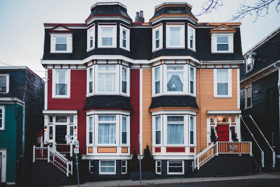 A three-storey Victorian-style house with a facade painted in vibrant red, orange, and cream colours, featuring multiple bay windows with white frames and curtains inside. The property has a dark grey roof with dormer windows, and the front entrance is accessible via a small set of white wooden stairs with a matching railing, leading to a red door decorated with a festive wreath. In front of the house, on the pavement, there are two small evergreen trees, a street sign, and a parked vehicle partially visible. The overall scene suggests preparation for a home relocation or a furniture transport process, with the house being part of a residential area. This image demonstrates typical stair access considerations for movers, consistent with house removals services by Man with Van St Johns.