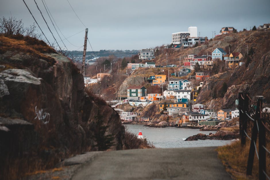 The image shows a narrow paved pathway leading towards a body of water with a buoy, situated at the base of a hillside. On the hillside, there are numerous residential houses of various sizes and colors, including blue, white, yellow, and red, with some featuring large windows and balconies. A few houses are positioned close to the water’s edge, while others are built at higher levels on the rocky slope. To the left, a steep rocky cliff faces the pathway, with visible chalk markings, and power lines supported by tall wooden poles run parallel to the path, extending into the distance. The scene is captured under overcast skies, with soft, diffused lighting highlighting the texture of the rocks, houses, and water. This setting is characteristic of a coastal home relocation area, where careful transportation and logistical planning are paramount for moving furniture and household items between hillside residences and the waterfront, as managed by companies like Man with Van St Johns.