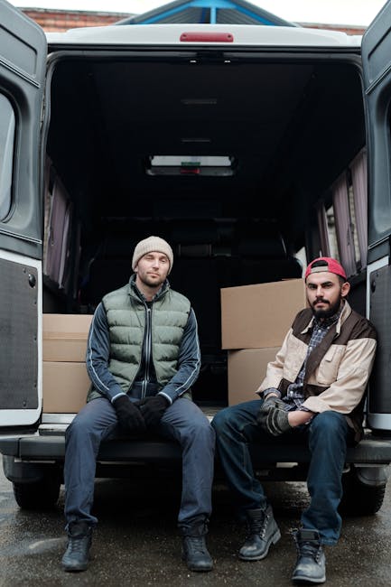 A grey moving van positioned on an unpaved area on a hill, with its side and rear doors open, revealing a wooden-framed mirror inside. The van is partially loaded with packaging materials, including c