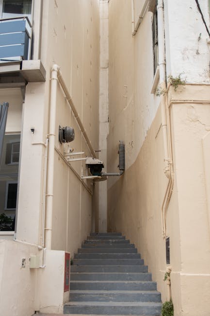 A narrow outdoor staircase with dark grey steps ascending between two tall, beige-painted walls of a building. The staircase is flanked by exposed white pipes and electrical meters attached to the walls, with some pipes running vertically and others horizontally. A small section of green foliage is visible at the bottom right corner near the base of the stairs. The walls show signs of weathering and age, with slight discoloration and cracks. This setting appears to be part of a residential or apartment complex in St Johns, where the challenges of moving furniture or boxes up a tight staircase may require professional removal services such as those provided by Man with Van St Johns, especially during a home relocation or furniture transport process.