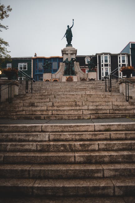 Photograph of the top view of a wide stone staircase leading up to a public monument featuring a statue of a woman holding an object in her raised hand, with additional statues at the base of the main sculpture. The staircase has metal railings on both sides and is situated outside in an urban setting, with residential buildings and trees visible behind the monument. The sky appears overcast, giving a grey tone to the scene. The image emphasizes the staircase's upward perspective, suitable for illustrating home relocation or moving services focused on navigating narrow Victorian staircases, as seen in St Johns flats, with potential involvement of furniture transport and loading processes handled by companies like Man with Van St Johns.