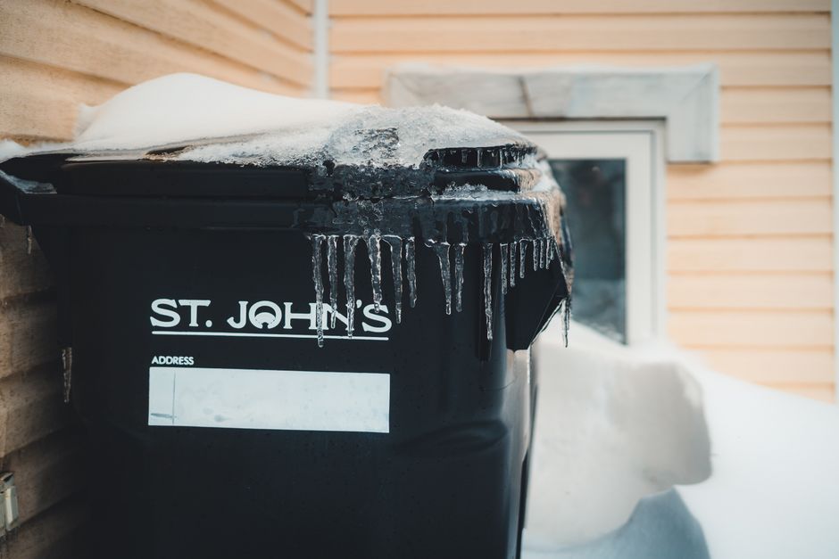 Close-up image of a black wheelie bin labeled 'ST. JOHN'S' located outside a residential property during winter, with snow accumulated on top and ice forming around the edges. The bin is positioned near a brick wall and a window with a white frame, which is partially visible in the background. The scene is outdoors, with natural daylight illuminating the area. This image illustrates typical waste disposal arrangements outside a home, relevant to house removals and moving services provided by Man with Van St Johns, especially in relation to preparing a property for a home relocation or moving process.