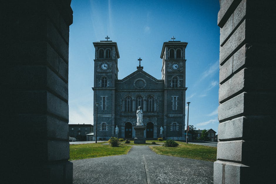 The image shows a view of a large stone church with twin towers, each topped with a cross, and a central statue of a religious figure in front of the building. The church's facade features arched windows and a clock on each tower, with detailed architectural stonework. The photo is taken from a shaded area between two brick walls, framing the church in the distance. The pathway leading up to the church is lined with small bushes and well-maintained grass, illuminated by daylight under a partly cloudy sky. This scene likely captures a moment during daylight hours of a church or religious building, with no visible moving or packing equipment. The image contextually supports themes of house removals or home relocation, as it emphasizes careful site access and the importance of planning for large-item transport and logistical considerations when moving or transporting large items, in line with services offered by Man with Van St Johns.