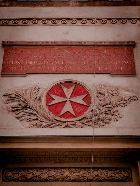 The image shows an ornate wall detail, featuring a carved emblem with a red circular background and a white cross at its center, surrounded by carved olive and laurel branches. Above the emblem, there is a rectangular stone plaque with Latin inscriptions, mounted on a smooth, light-colored wall. The section of the wall displays decorative molding and a horizontal band with classical motifs. The scene appears to be indoors, with soft ambient lighting highlighting the textured carvings. In a house removal context, this detail could be part of a property's interior wall during a moving or packing process by Man with Van St Johns, indicating careful handling of valuable or period-specific architectural features as part of a professional relocation service.