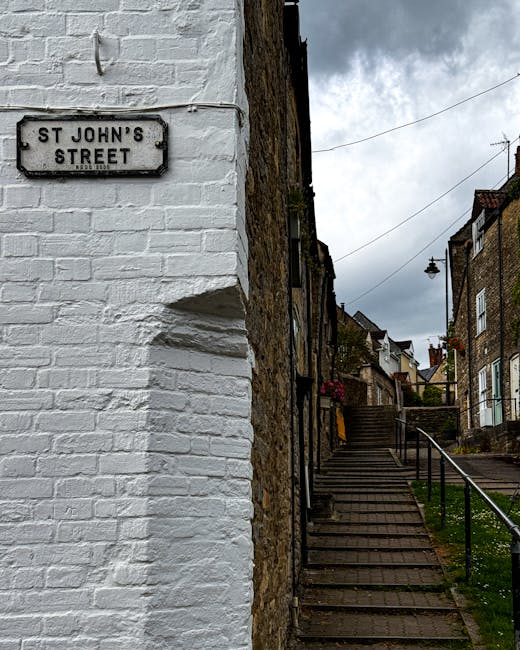 A close-up view of a white-painted brick corner of a building on St John's Street, with a black and white street sign mounted on the wall indicating the street name. In the background, a steep outdoor staircase made of wooden steps ascends alongside a stone retaining wall, leading up to residential houses with brick facades and garden decorations, such as flower baskets and potted plants. A metal railing runs parallel to the stairs for support, and a streetlamp hangs from a wire above the pathway. The sky is overcast with grey clouds, suggesting typical weather during a home relocation process. The scene captures an urban residential area likely used by Man with Van St Johns during furniture transport and packing operations for house removals, emphasizing the staircase access and street environment involved in local moving services.