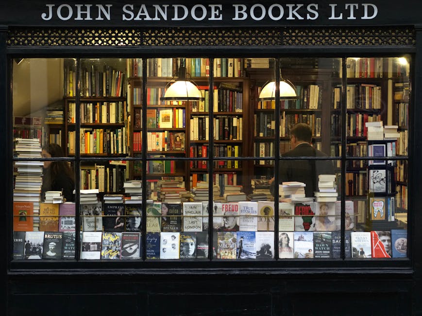 View through the window of John Sandoe Books Ltd, displaying neatly arranged wooden bookshelves filled with a diverse selection of hardcover and paperback books in various sizes and colours, some stacked horizontally and others standing vertically. Two hanging pendant lamps with warm lighting illuminate the interior, highlighting the dense arrangement of literary titles. Inside the shop, two individuals are visible, one on the left and one on the right, browsing or working among the shelves. The window display includes a variety of books laid flat on the lower ledge, facing outward for easy viewing, with some standing upright side-by-side. The exterior features a black framework with the shop name in white above the display window. This scene depicts a quiet, well-organized bookselling environment, suitable for home relocation or packing and moving services focusing on specialist bookstores or interior shelving arrangements.