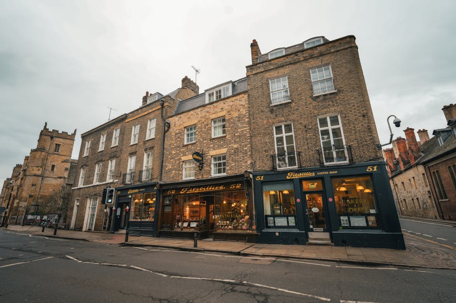 The image depicts a row of historic brick buildings on St Johns High Street, featuring a ground-floor bakery and coffee shop with large glass windows displaying baked goods and a menu, illuminated by interior lighting. The upper floors are residential, with multiple white-framed sash windows, some with small balconies featuring black wrought iron railings. The sky is overcast, casting soft natural light over the scene, with a quiet street in front showing a few parked bicycles and bollards along the pavement. The bakery signs are in gold lettering, and the building facade includes traditional elements such as brickwork and chimney stacks. This setting illustrates a typical street scene suitable for house removals, home relocation, or furniture transport services, as offered by Man with Van St Johns, highlighting the environment where professional packing, loading, and transport logistics might be involved in a narrow shopfront urban context.