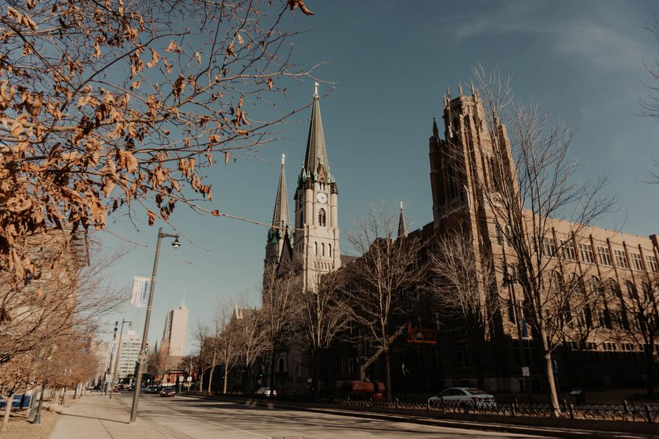 A street scene in St Johns featuring a large historic church with two tall spires and a clock on one of the spires, surrounded by leafless trees and modern buildings. The sky is clear and blue, with sunlight casting shadows on the pavement and the church. The street has parked cars and a few moving vehicles, and a lamppost with a banner attached. This urban environment depicts an area where home relocation and furniture transport services, like those provided by Man with Van St Johns, may support residents with removals and packing during house moves or local furniture collection and delivery. The image captures the process of moving logistics, with the church situated in a central part of the town, providing context for property or furniture transport in a residential or commercial setting.